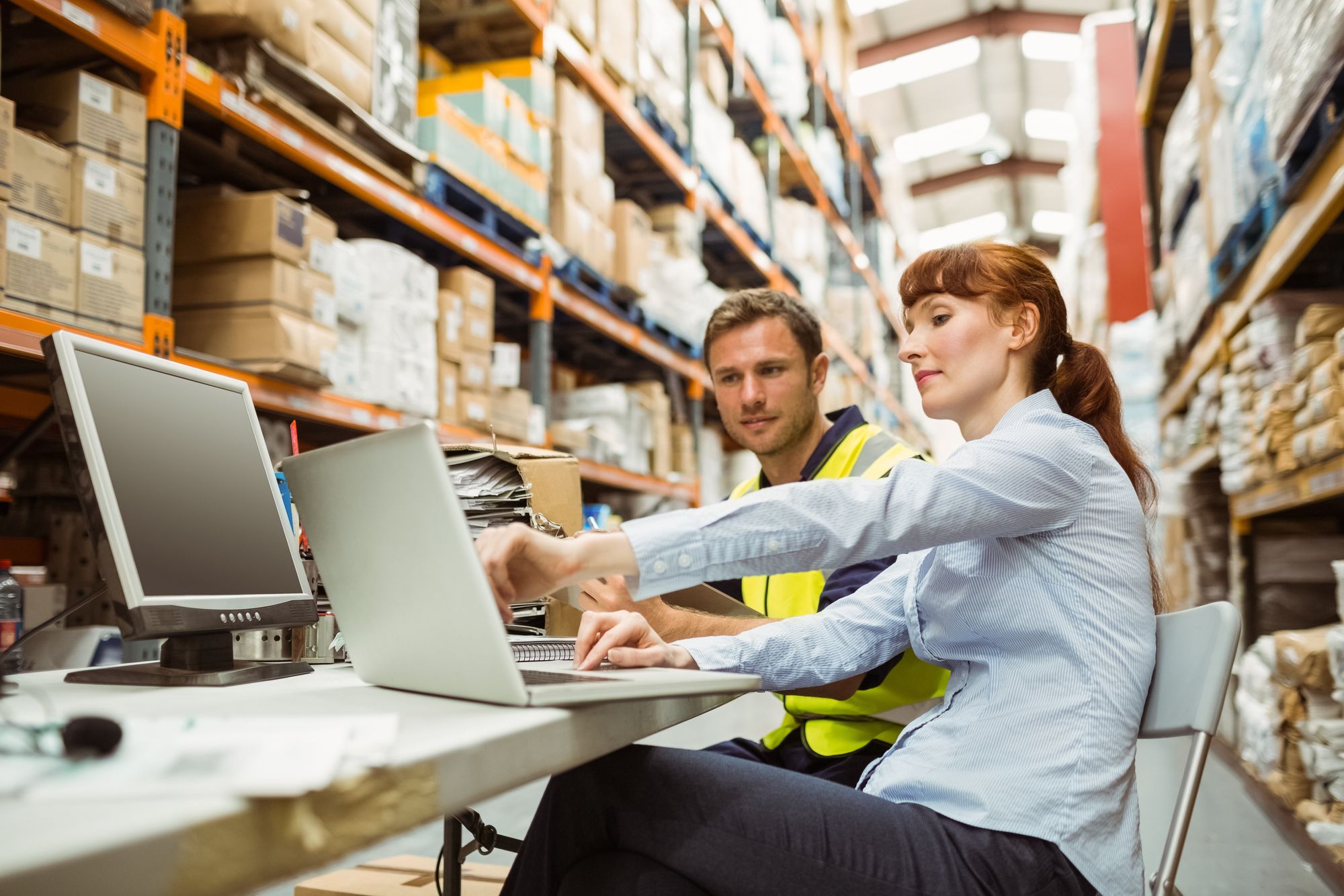 A woman showing a warehouse worker something on a laptop screen inside a warehouse